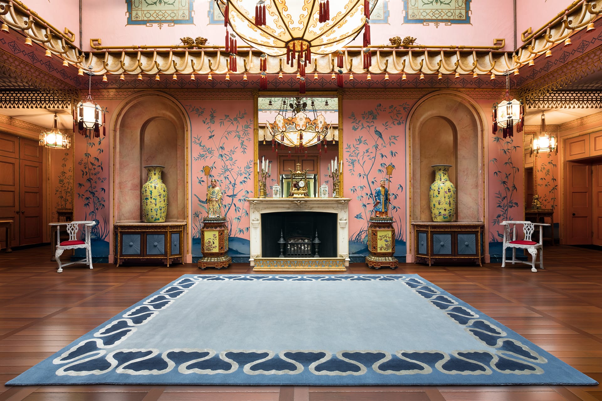 Ornate room with pink walls, floral patterns, large vases, chandelier, and blue rug centered on wooden floor.