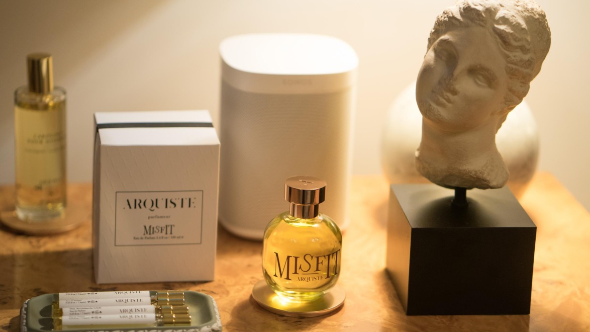 Perfume bottles and sculpted bust on a table next to a speaker, with a decorative box in the background.