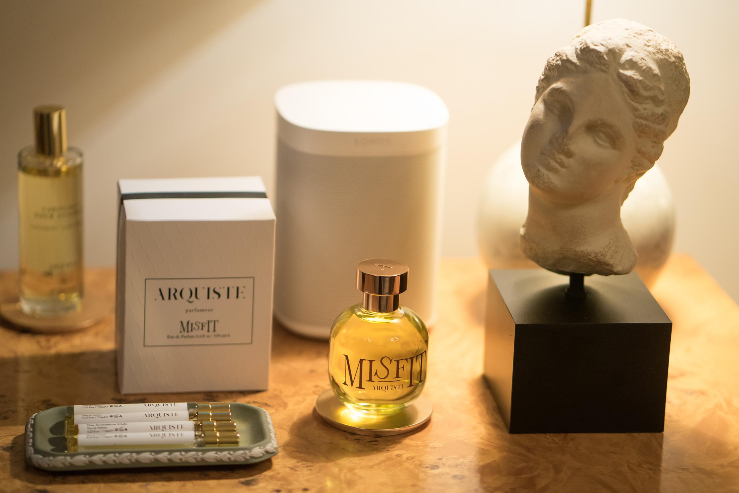 Perfume bottles and sculpted bust on a table next to a speaker, with a decorative box in the background.