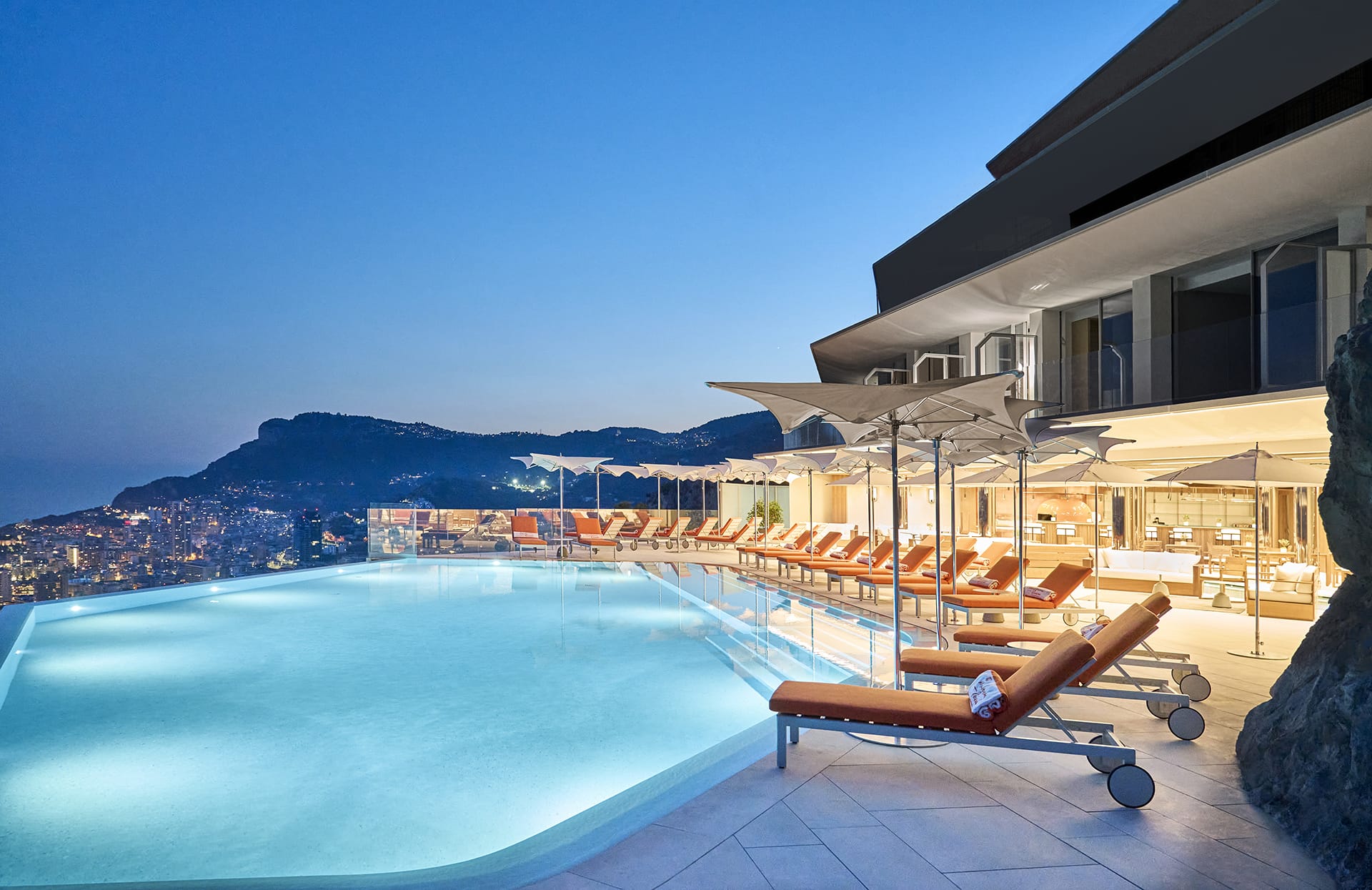 Infinity pool with lounge chairs overlooking a cityscape at dusk, surrounded by mountains and a modern building.