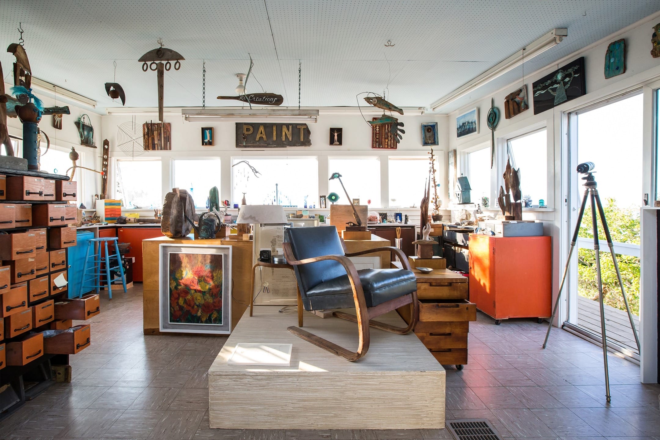 Artist's studio with various paintings, sculptures, colorful furniture, and natural light streaming in through large windows.