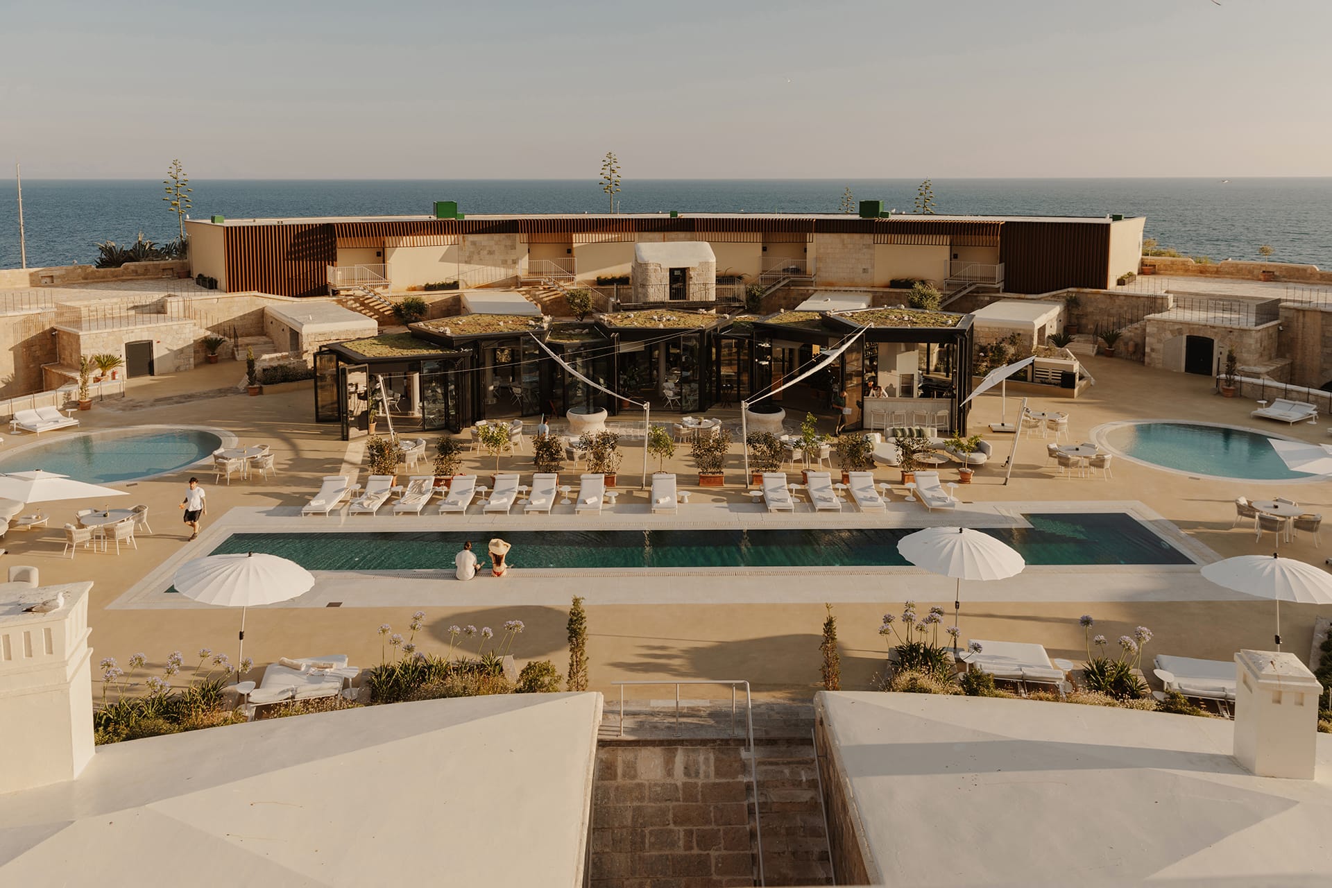 Luxury seaside resort with swimming pools, sun loungers, and umbrellas overlooking the ocean, showcasing modern architecture.