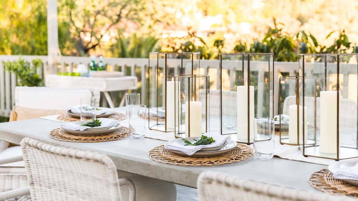 Outdoor dining table set with plates, napkins, and candles, surrounded by greenery and sunshine.