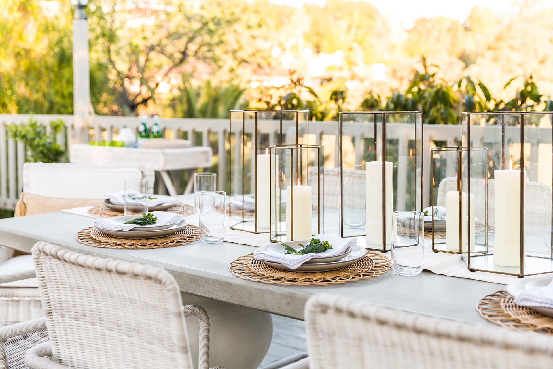 Outdoor dining table set with plates, napkins, and candles, surrounded by greenery and sunshine.