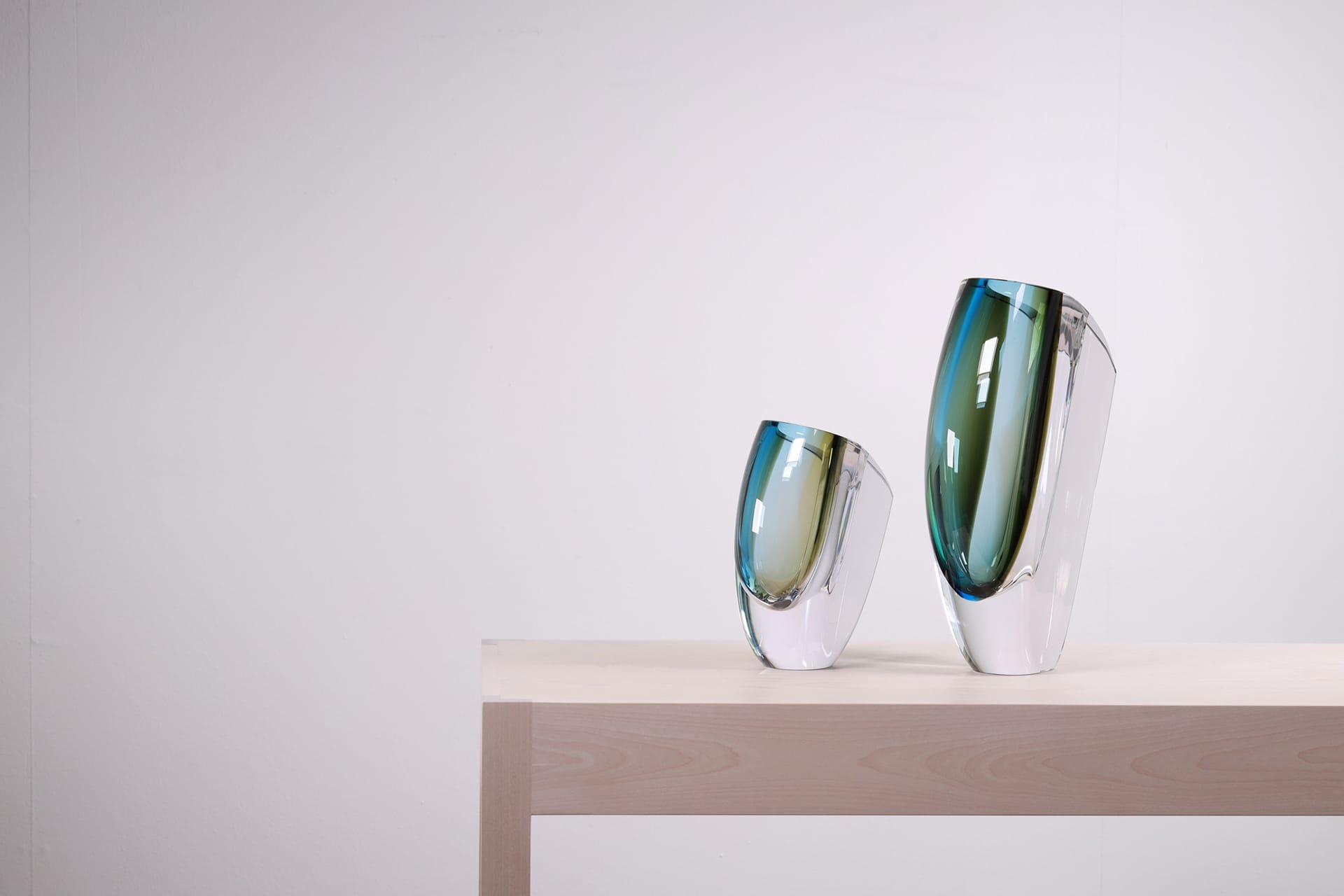 Two modern glass vases with blue and green tint on a light wooden table against a plain white background.