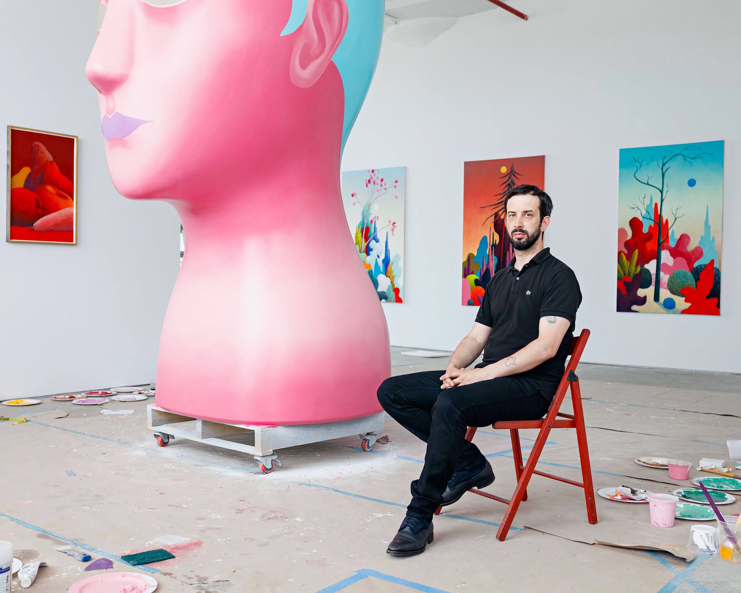 Artist sitting on a chair in a studio with vibrant paintings and a large pink sculpture in the background.