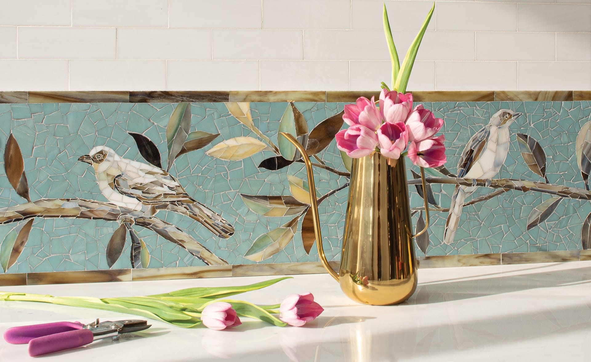 Golden vase with pink tulips on a counter in front of a bird mosaic wall.