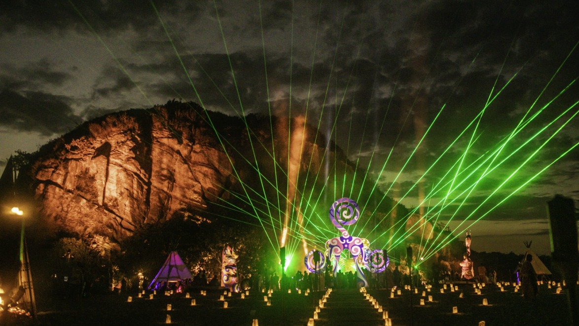 A nighttime festival scene with laser lights, illuminated rock formations, and glowing decorations in an outdoor setting.