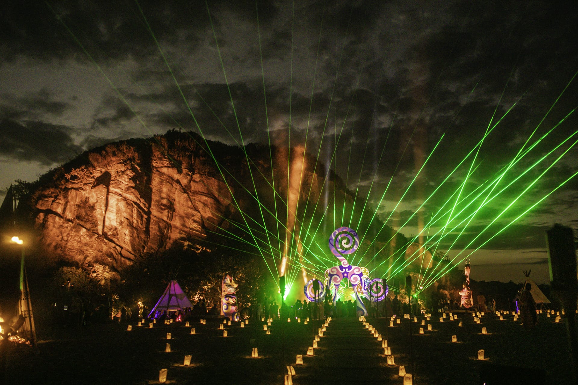 A nighttime festival scene with laser lights, illuminated rock formations, and glowing decorations in an outdoor setting.