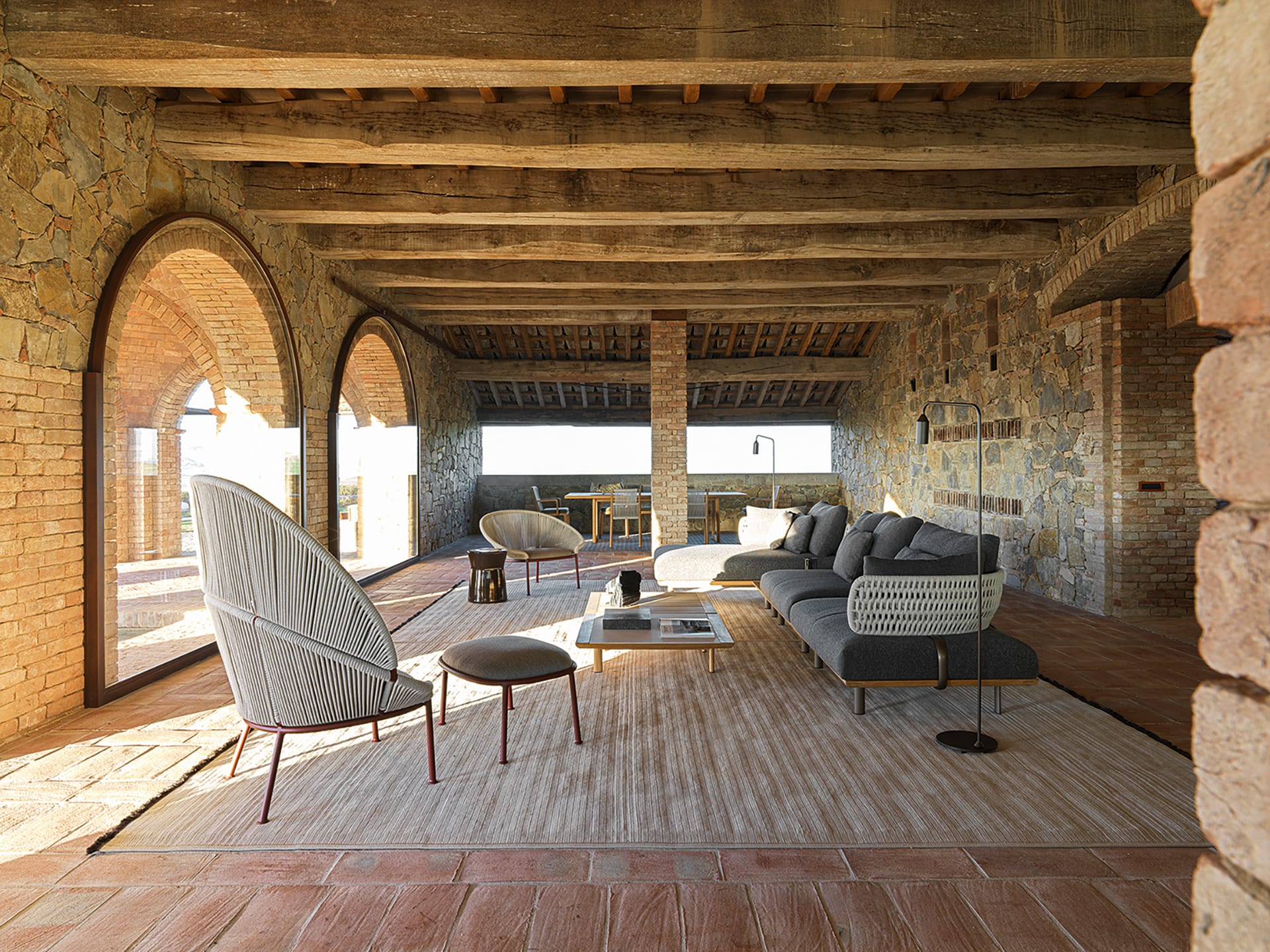 Rustic living room with stone walls, arched windows, wooden ceiling, modern furniture, and a large gray sofa.