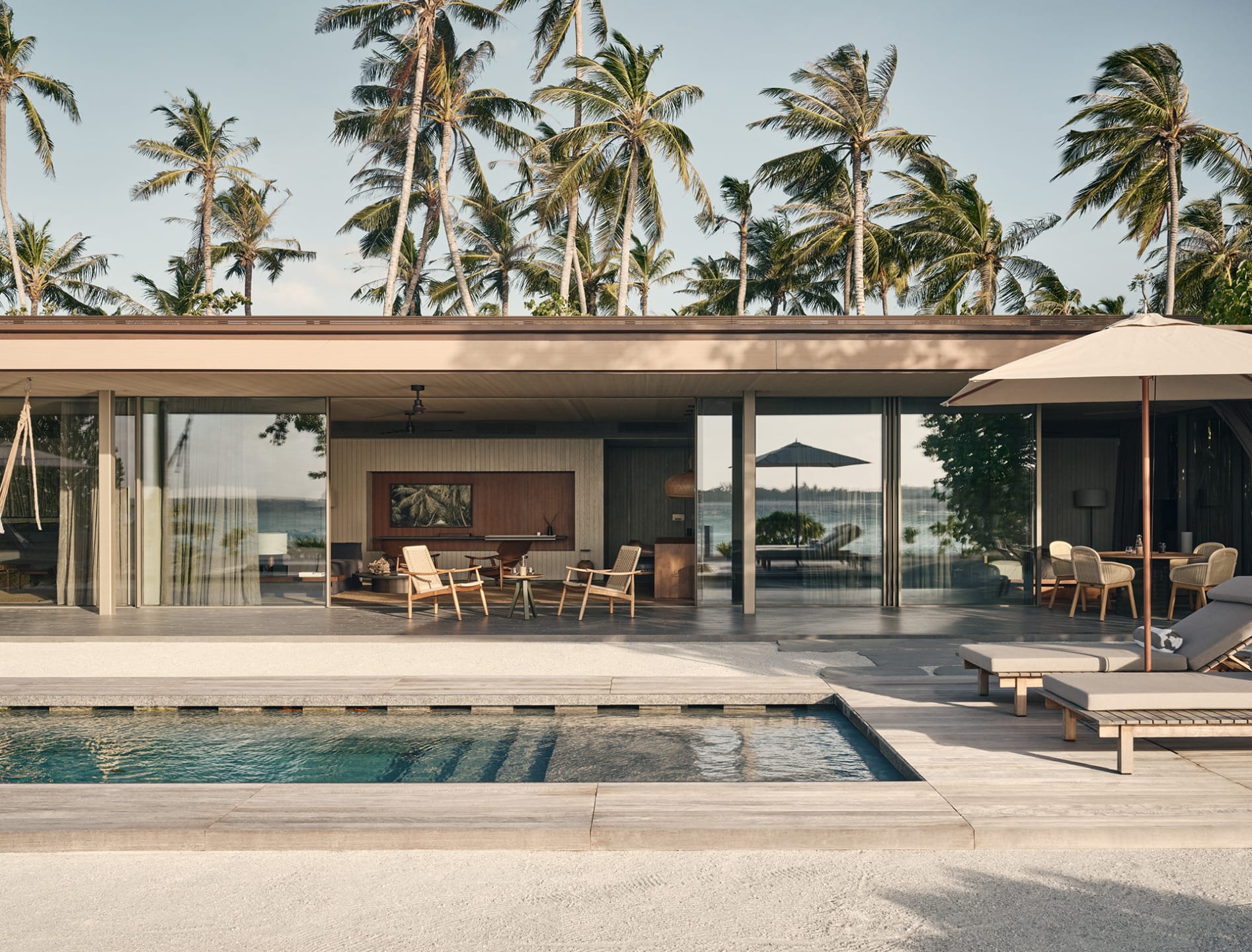 Modern beachfront villa with pool, palm trees in the background, lounge chairs, and outdoor dining area under umbrellas.
