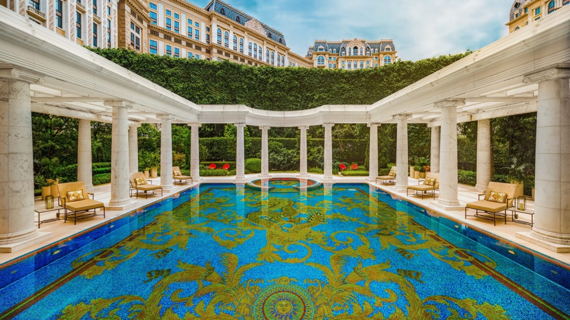 Luxurious hotel pool with ornate mosaic design, surrounded by elegant architecture and lush greenery under a clear blue sky.