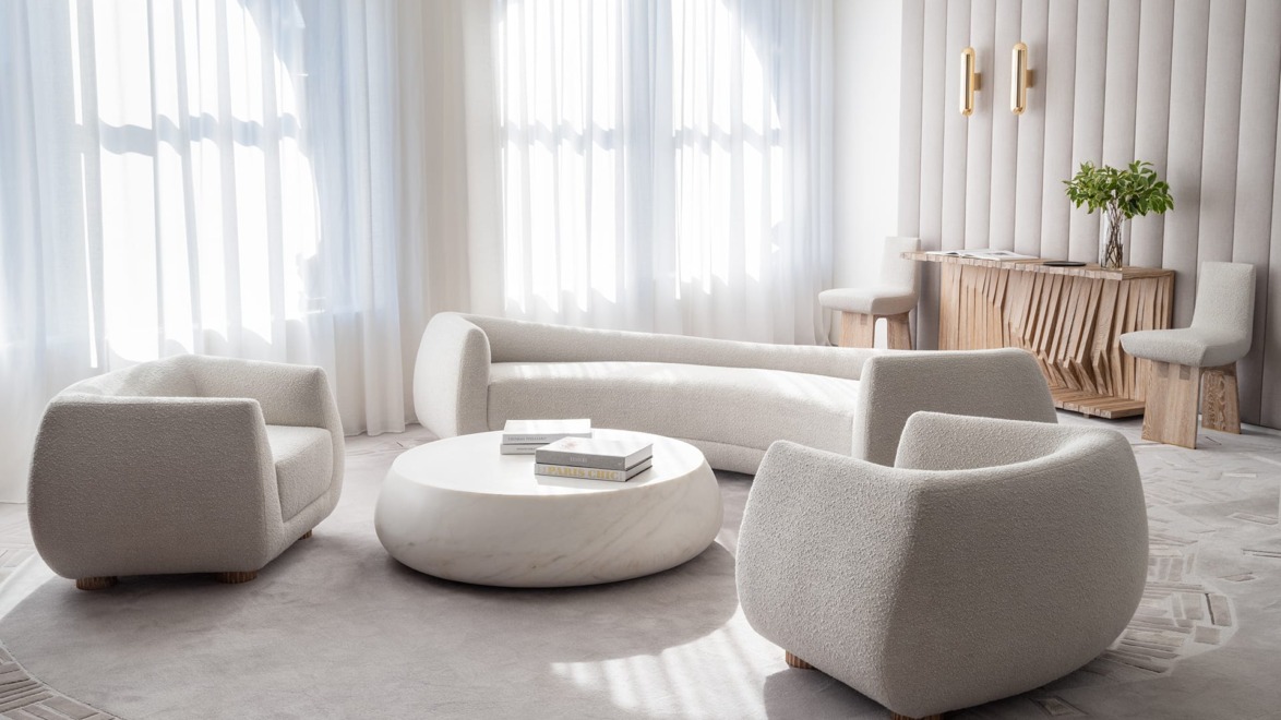 Modern minimalist living room with cream furniture, round marble table, and sunlight streaming through large windows.