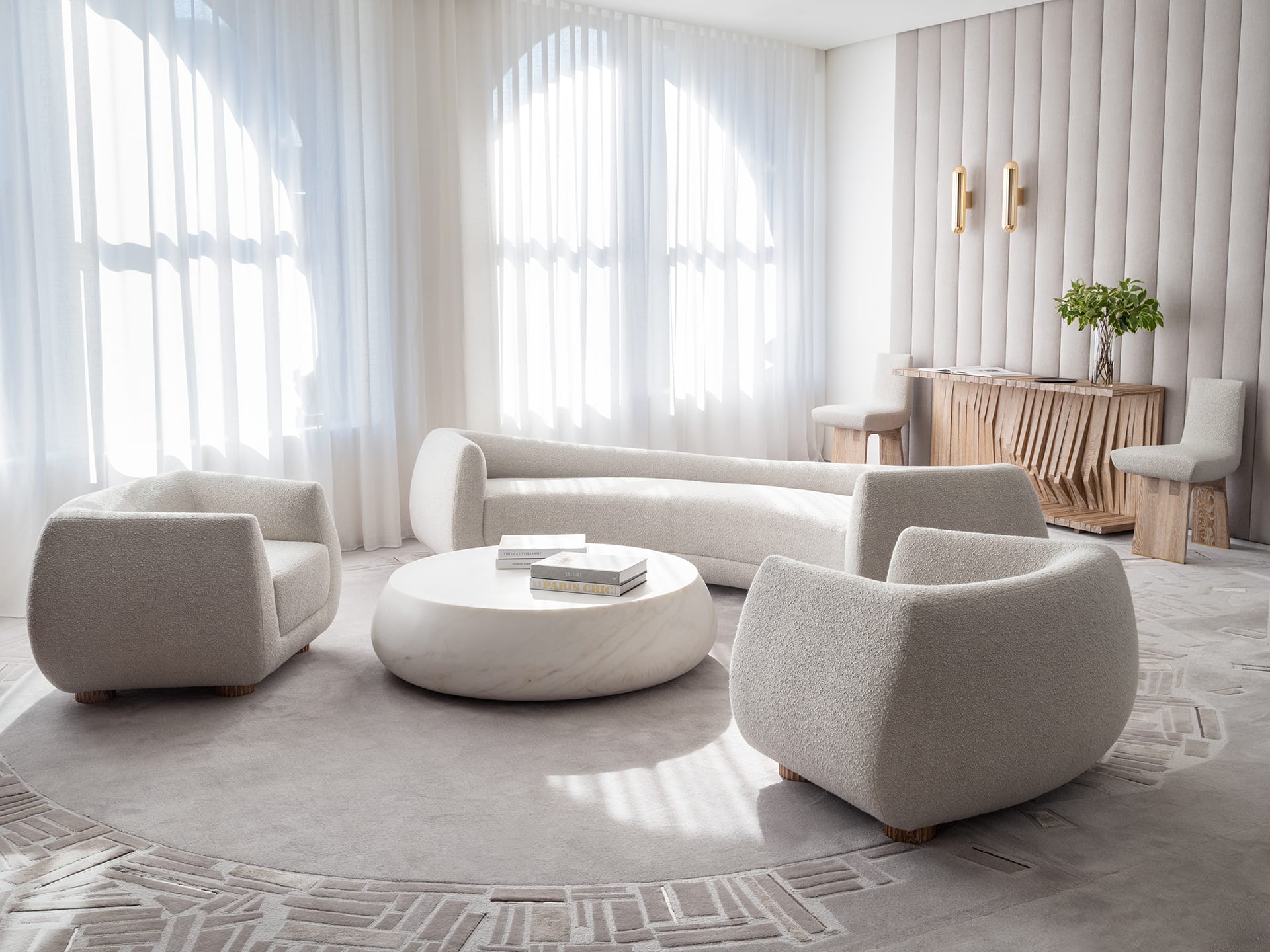 Modern minimalist living room with cream furniture, round marble table, and sunlight streaming through large windows.