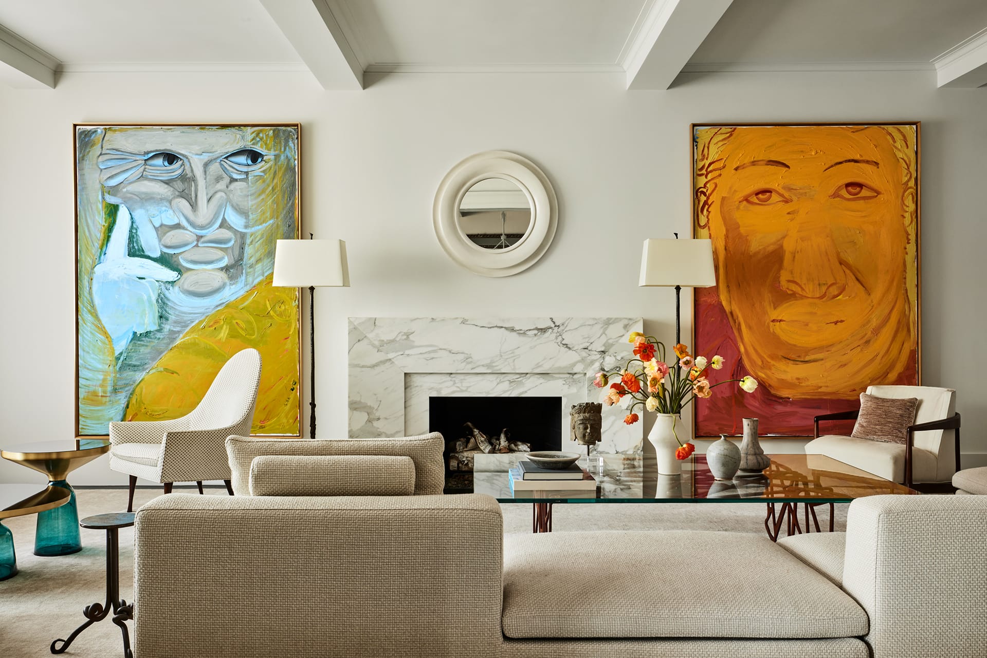 Modern living room with abstract art, beige sofas, marble fireplace, round mirror, and decorative table with flowers.