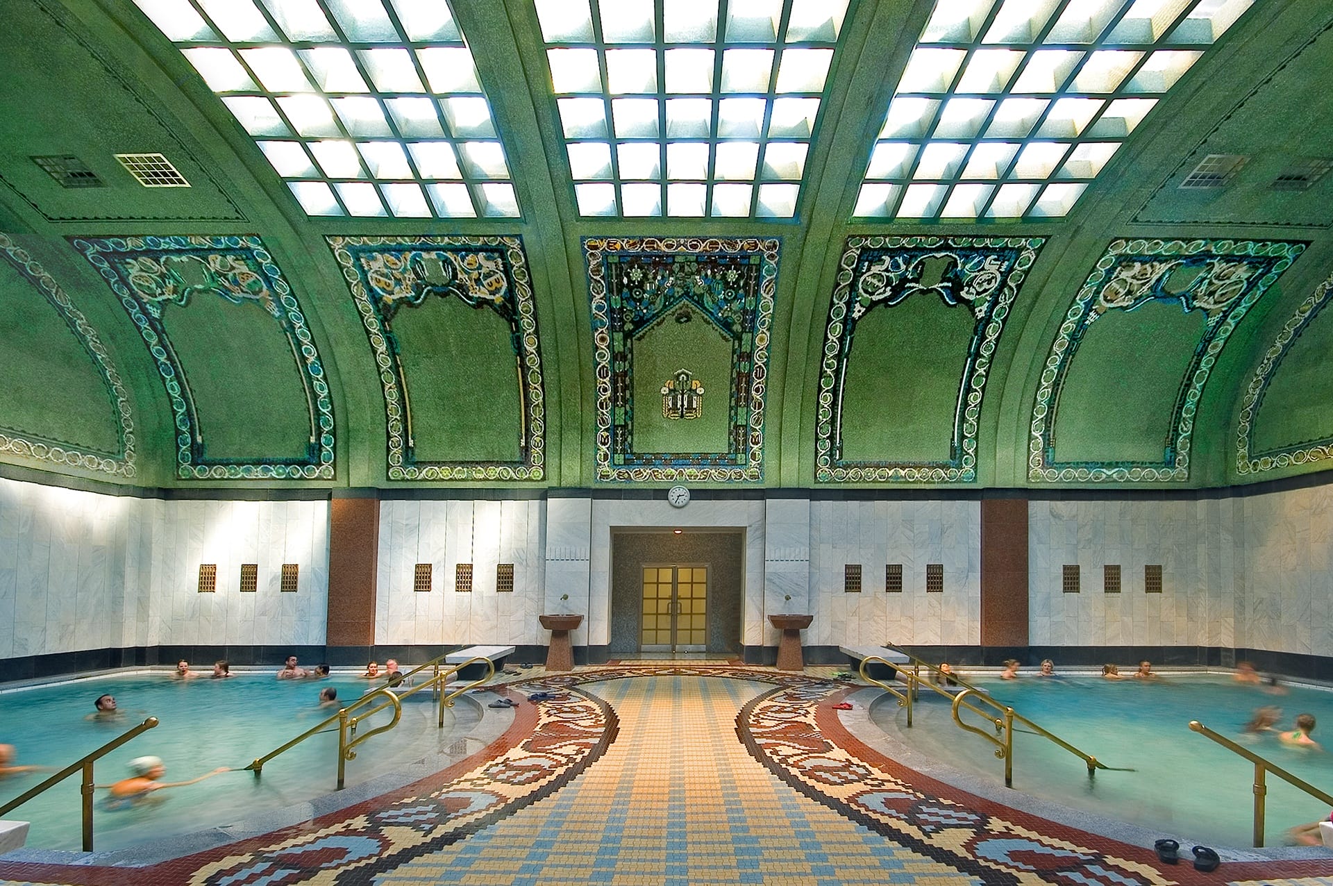 People relaxing in an ornate indoor swimming pool with mosaic tiles and a decorative arched ceiling.