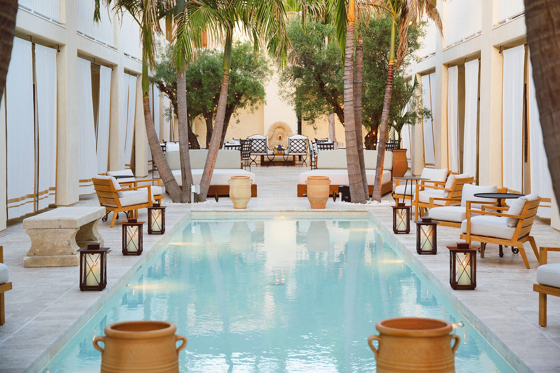 Luxury pool area with palm trees and elegant seating surrounded by lanterns and Mediterranean architecture.
