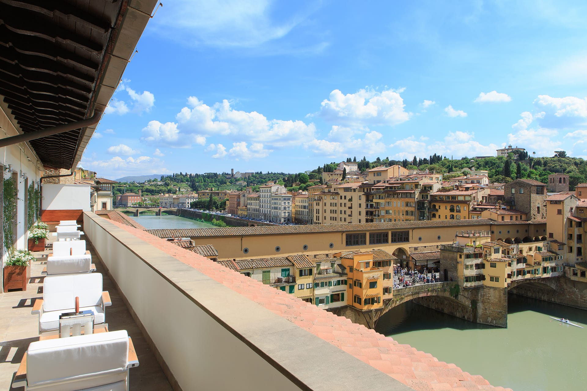 View of Ponte Vecchio and Arno River from a terrace with white chairs and tables under a sunny sky with scattered clouds.