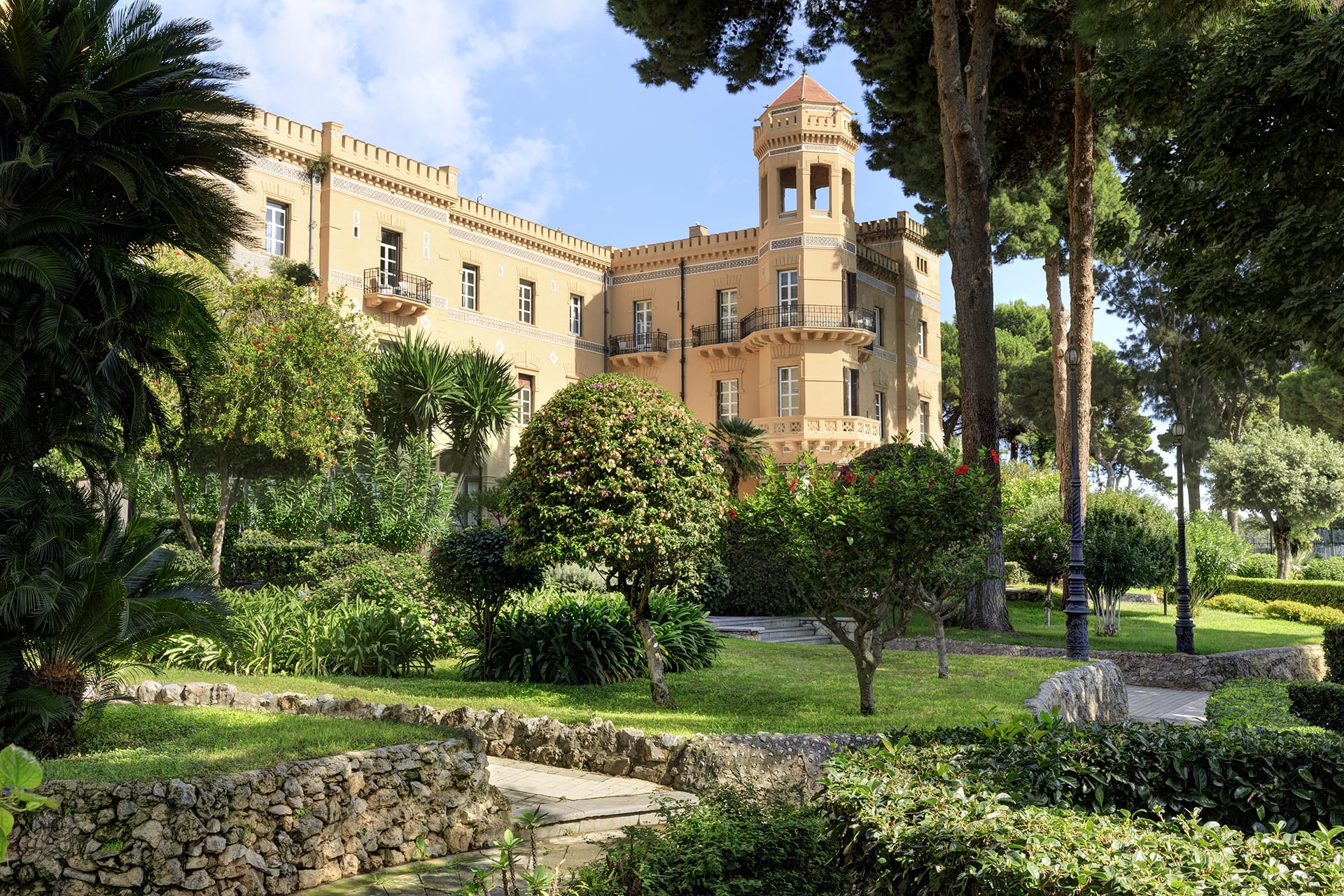 Historic building surrounded by lush gardens and tall trees with a stone pathway in the foreground under a blue sky.
