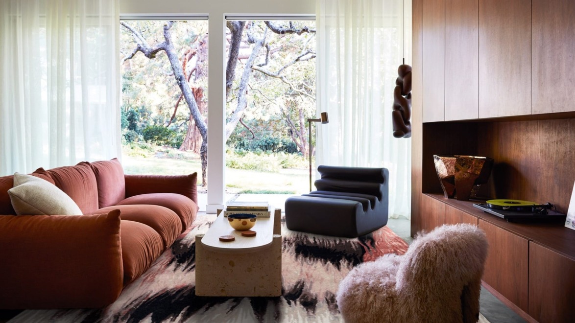 Modern living room with large windows, cozy sofas, abstract rug, and garden view outside. Stylish and inviting space.