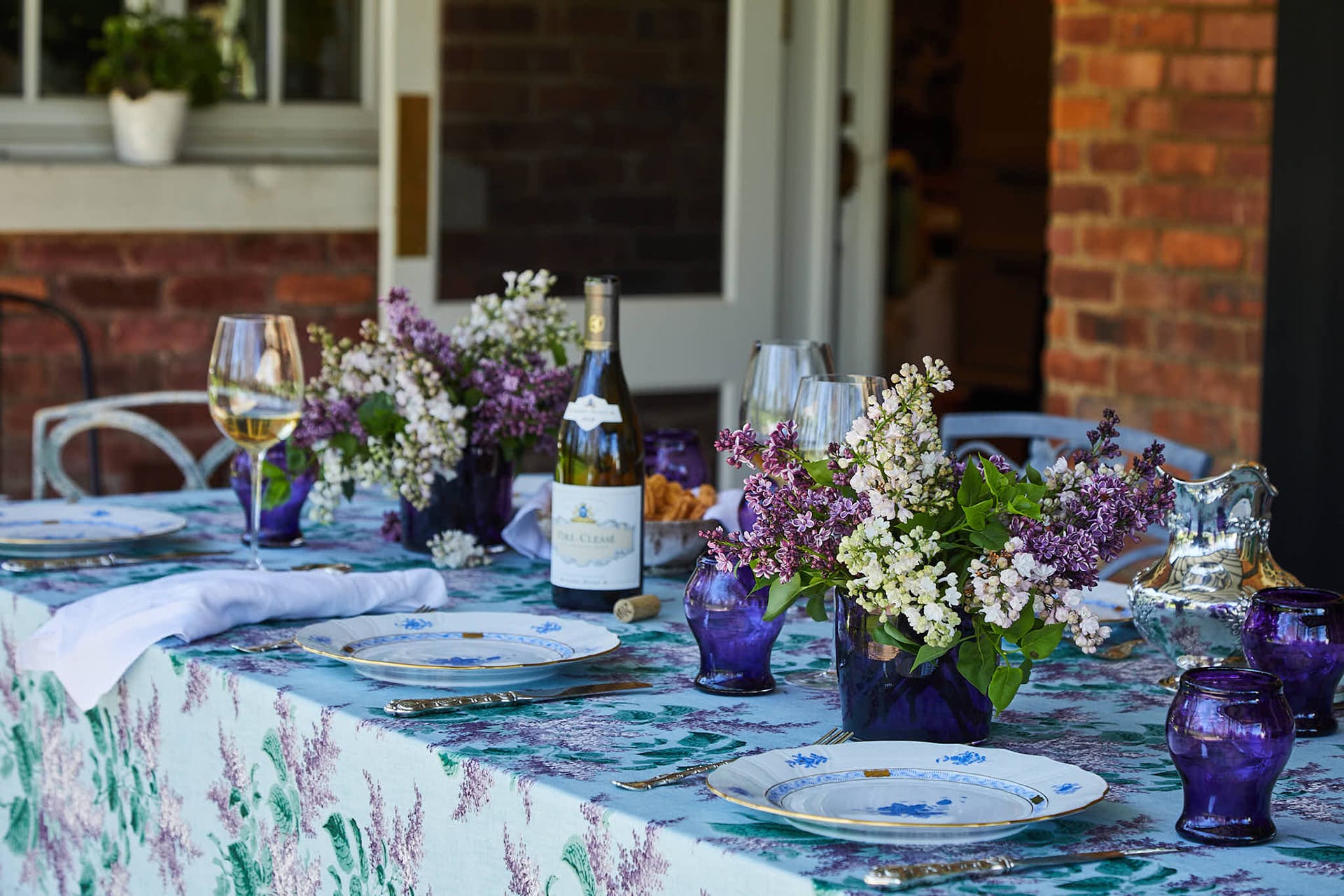 Outdoor dining table set with floral centerpiece, wine bottle, glasses, and plates on a patterned tablecloth in a garden setting.