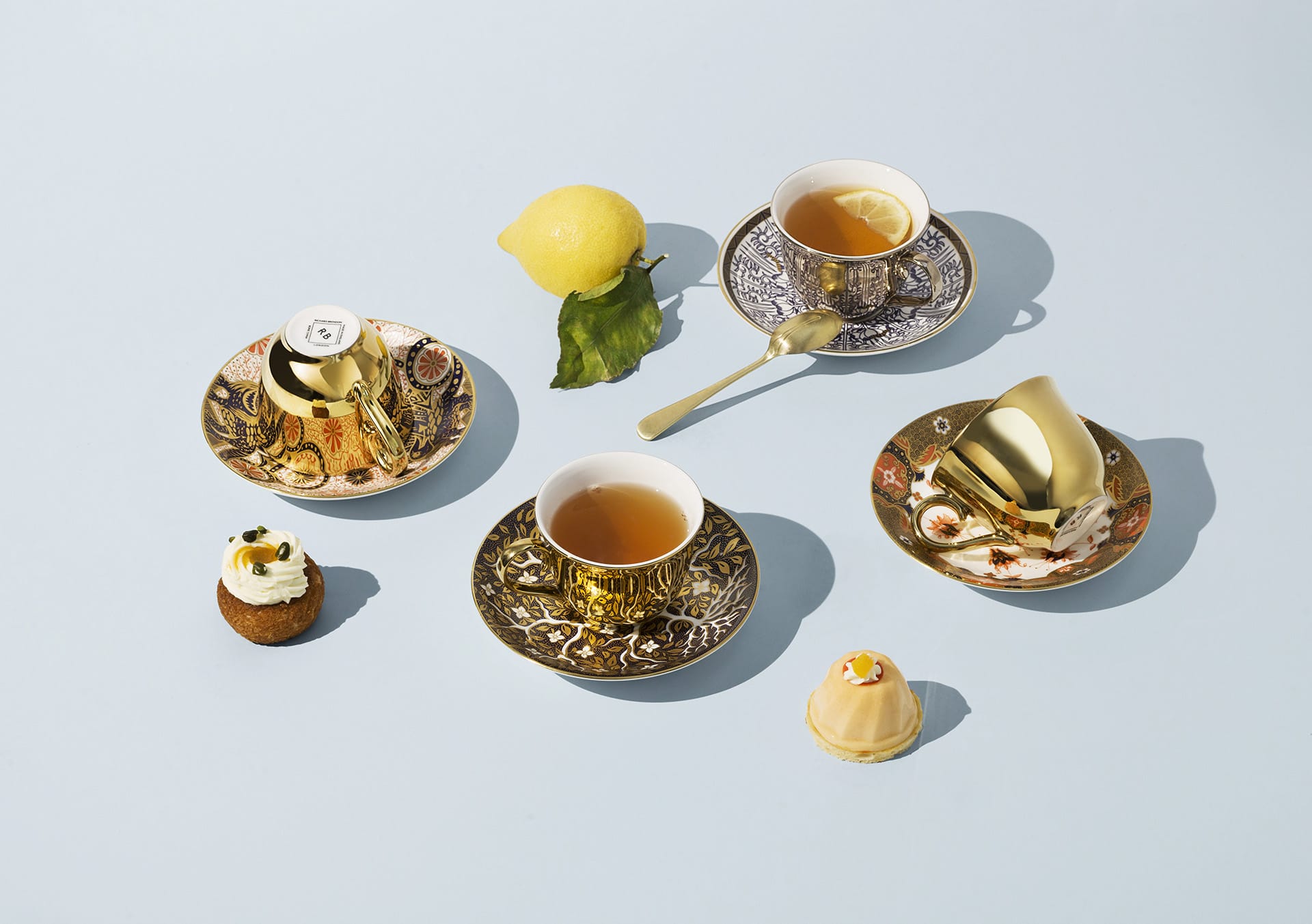 Decorative teacups and saucers with tea, lemon, and small pastries on a light blue background