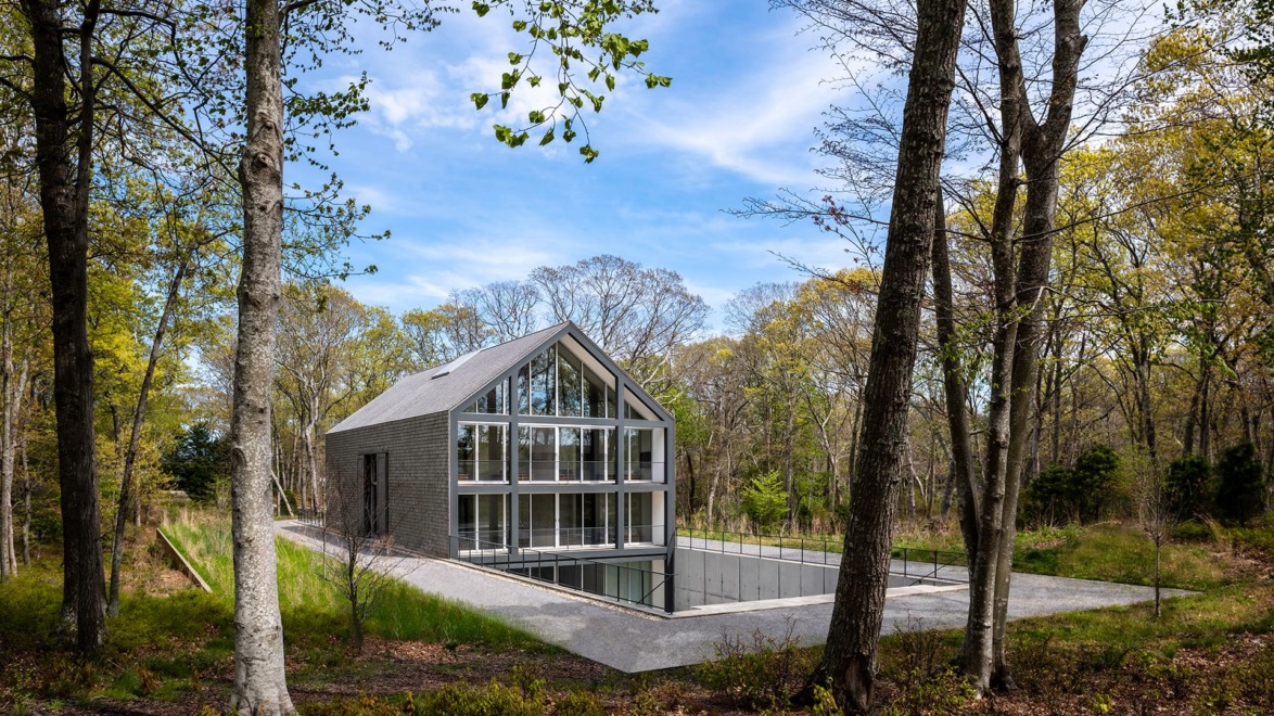 Modern glass and brick house surrounded by trees with a forest backdrop and open skies.