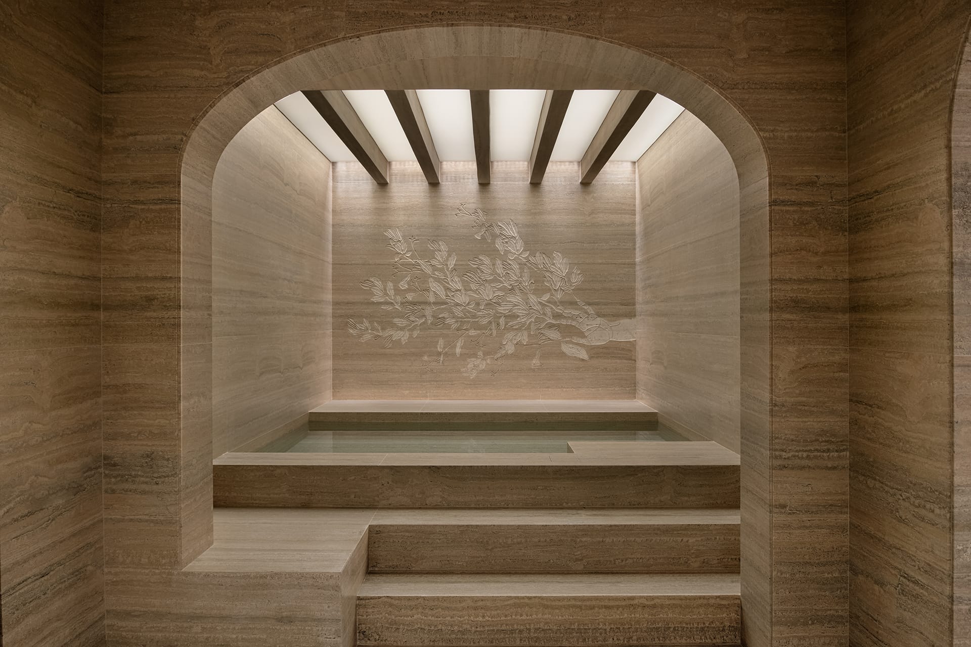 Luxurious travertine bath with intricate floral wall design and arched entry, featuring soft lighting and elegant steps