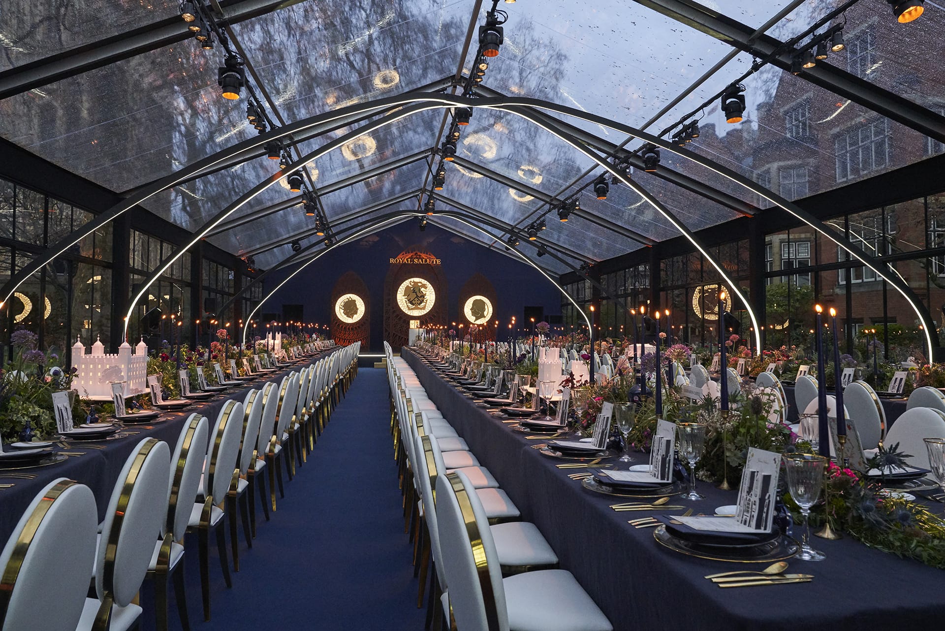 Elegant banquet setup under a clear tent with long tables, white chairs, green centerpieces, and circular lights overhead.