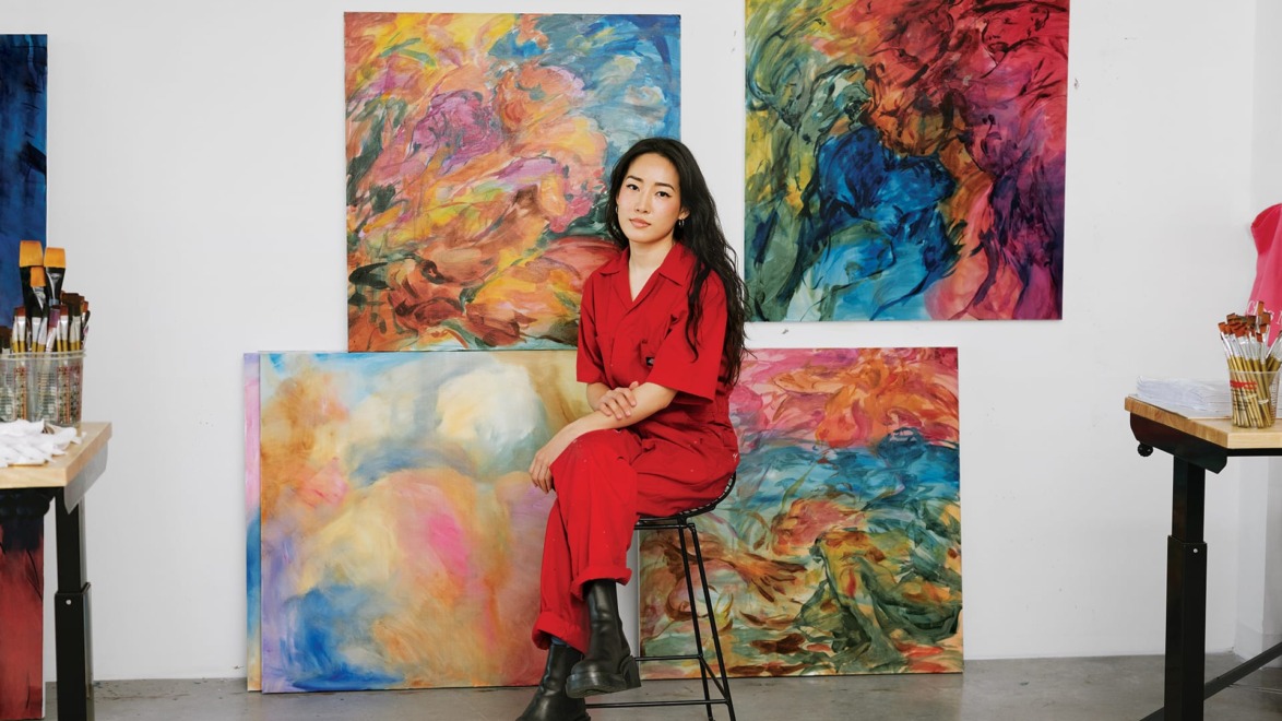 Artist in a red outfit sitting on a stool in front of vibrant abstract paintings in a studio.