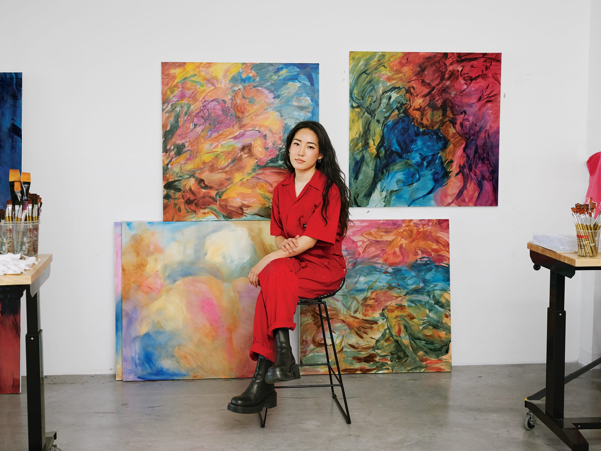 Artist in a red outfit sitting on a stool in front of vibrant abstract paintings in a studio.