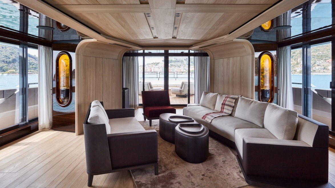 Luxurious yacht interior with modern furniture, wood paneling, and large windows overlooking a scenic view of water and mountains.