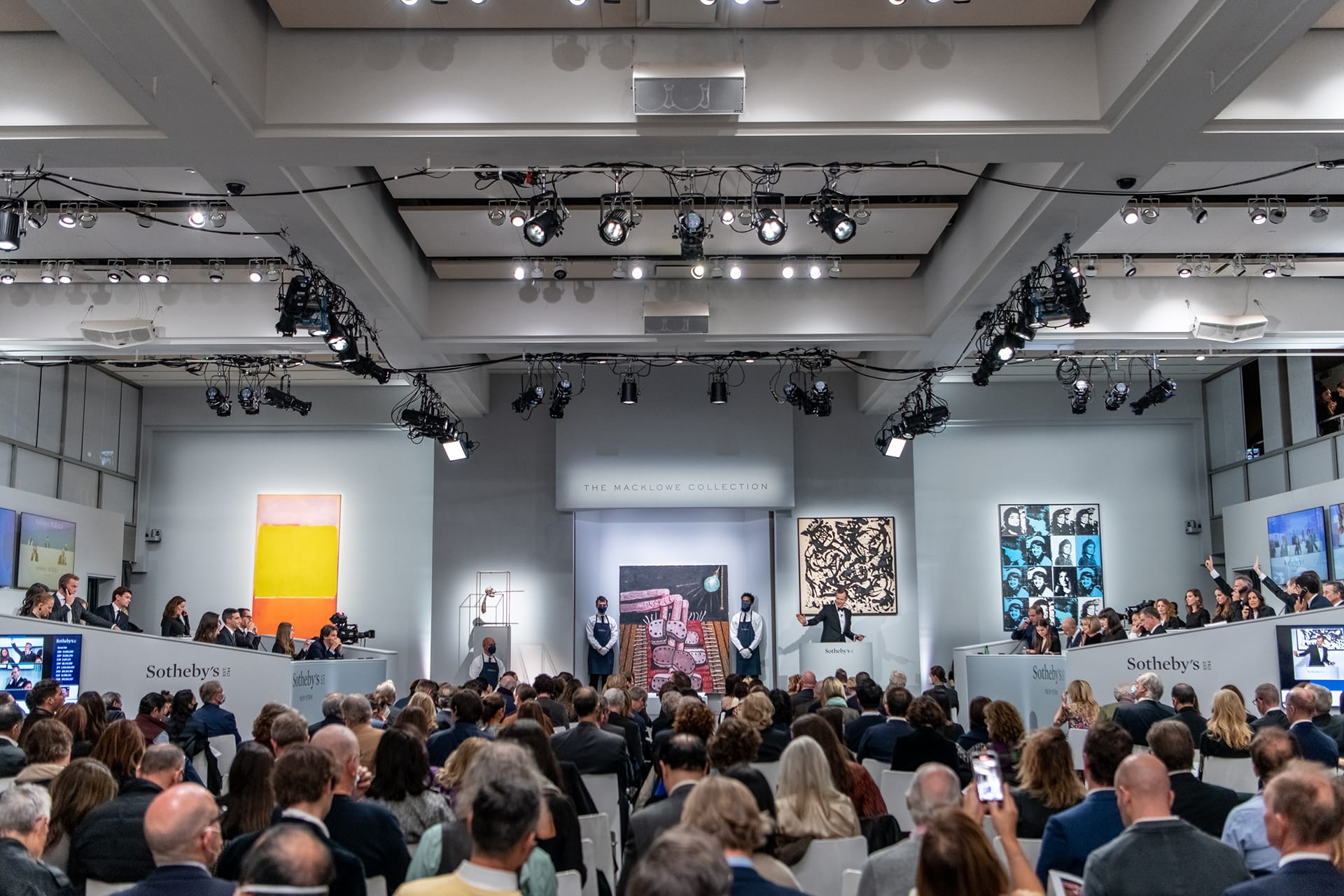 Art auction at a gallery with a large audience, paintings displayed, auctioneers standing at the podium.