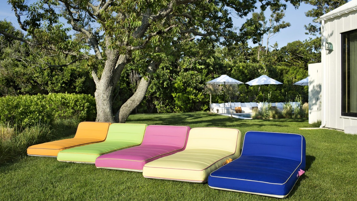 Colorful outdoor lounge chairs on grass with trees and umbrellas in the background on a sunny day.