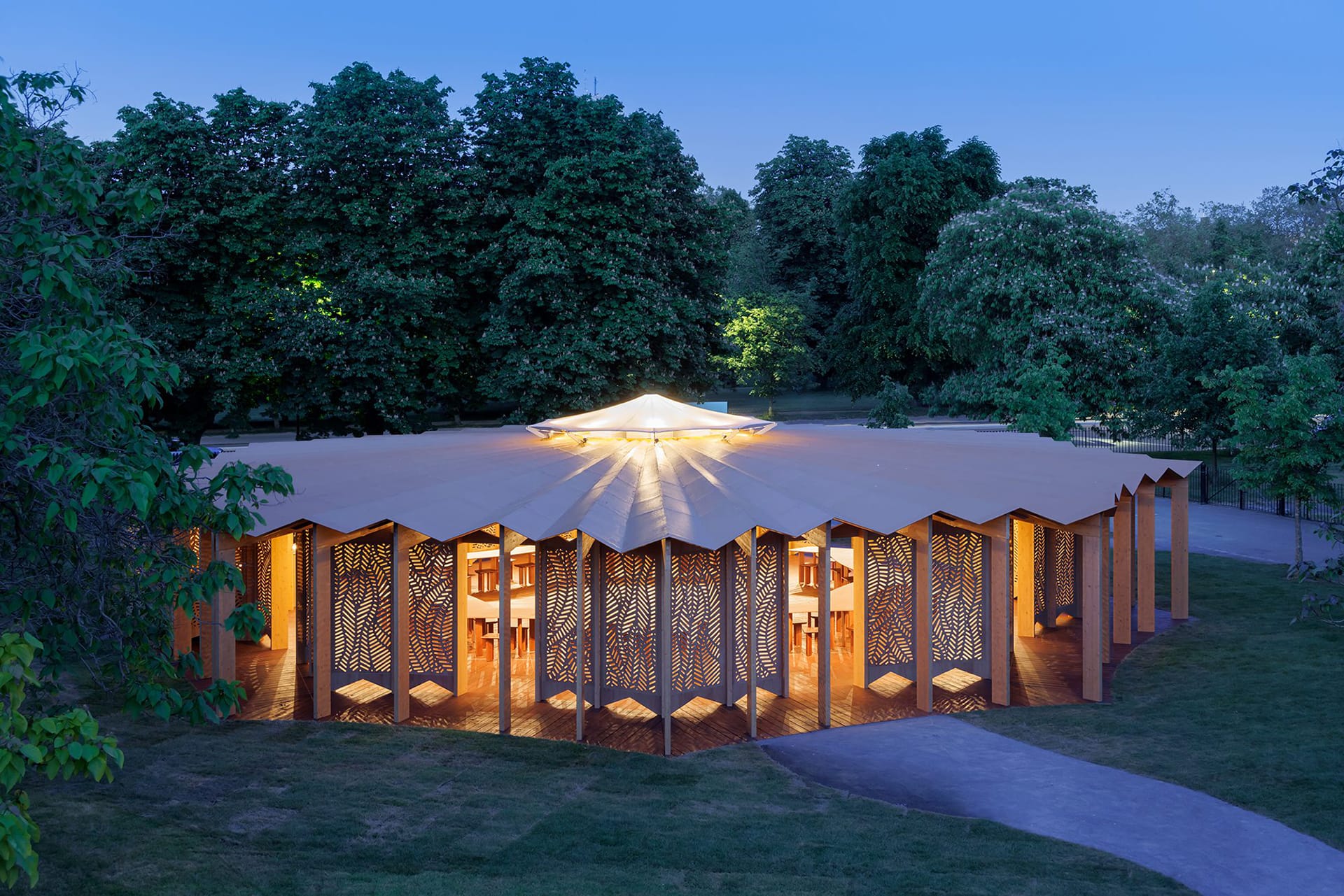 Modern pavilion with illuminated interior at dusk surrounded by lush trees in a park setting.