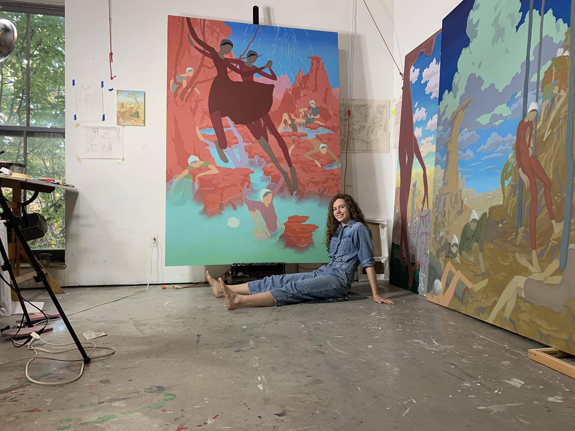 Artist sitting on studio floor, surrounded by colorful paintings, natural light streaming through large window.