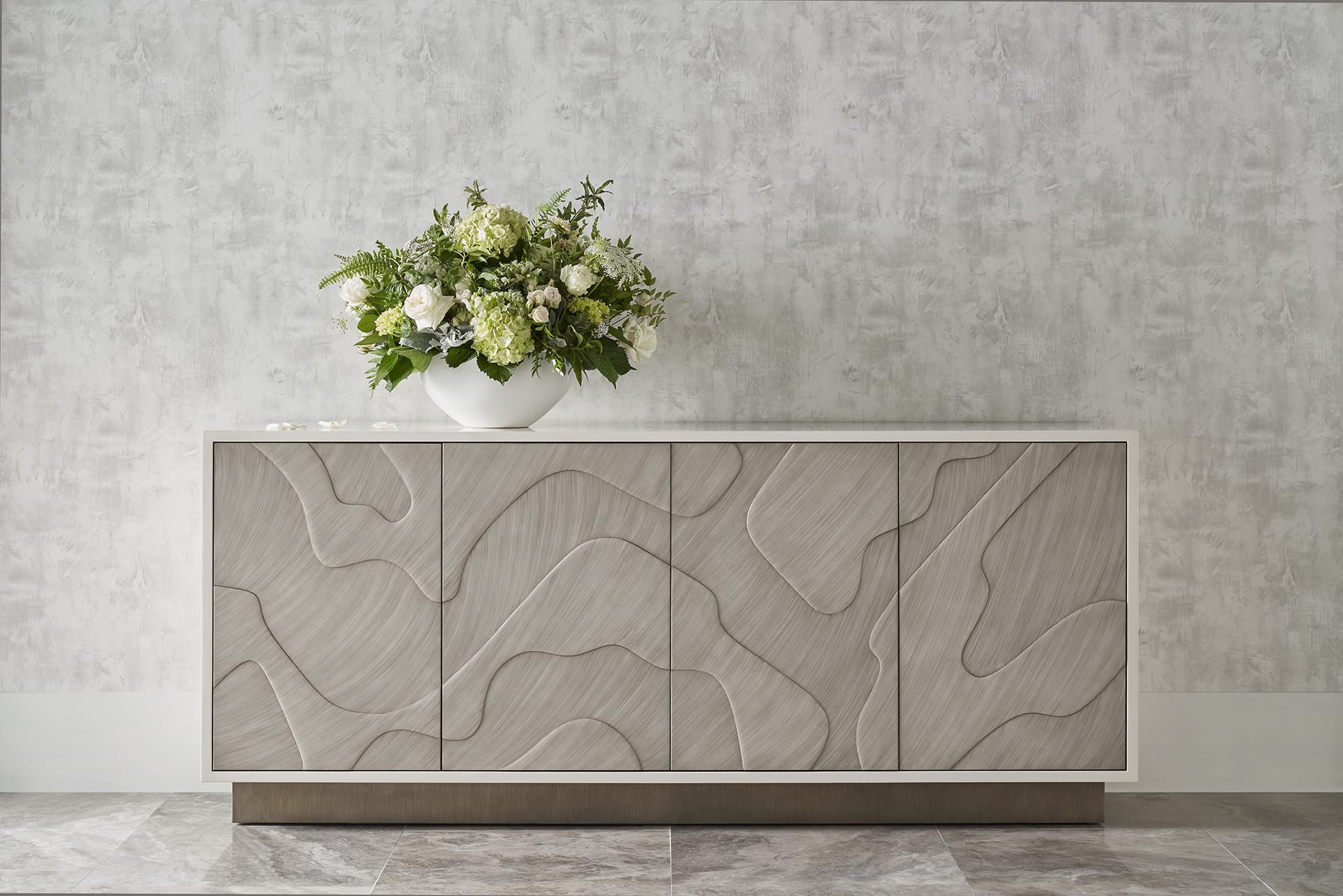 Modern sideboard with abstract design and a white vase of green and white flowers on top, against a textured wall.