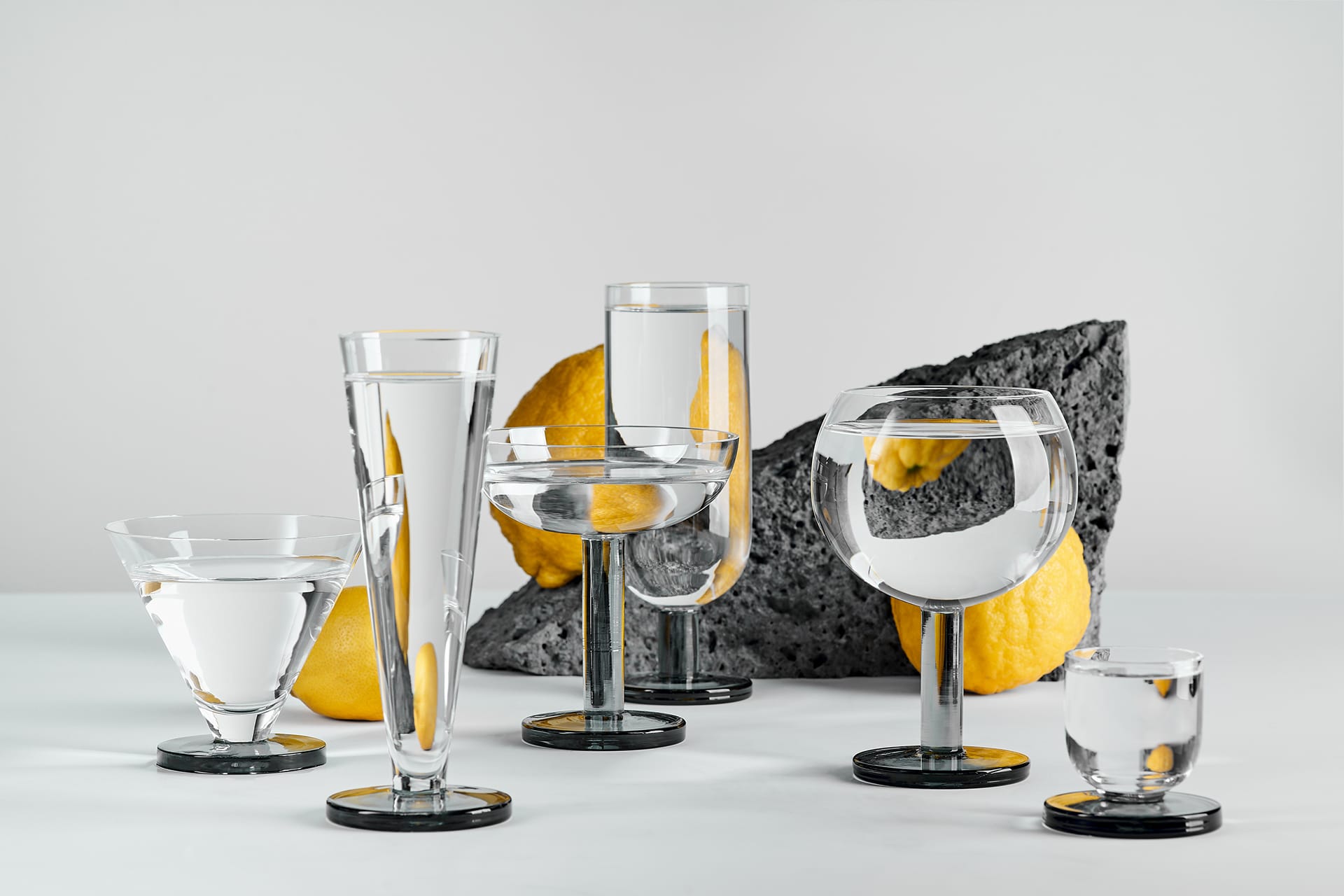 Assorted glassware filled with water in front of lemons and stone on a white background.