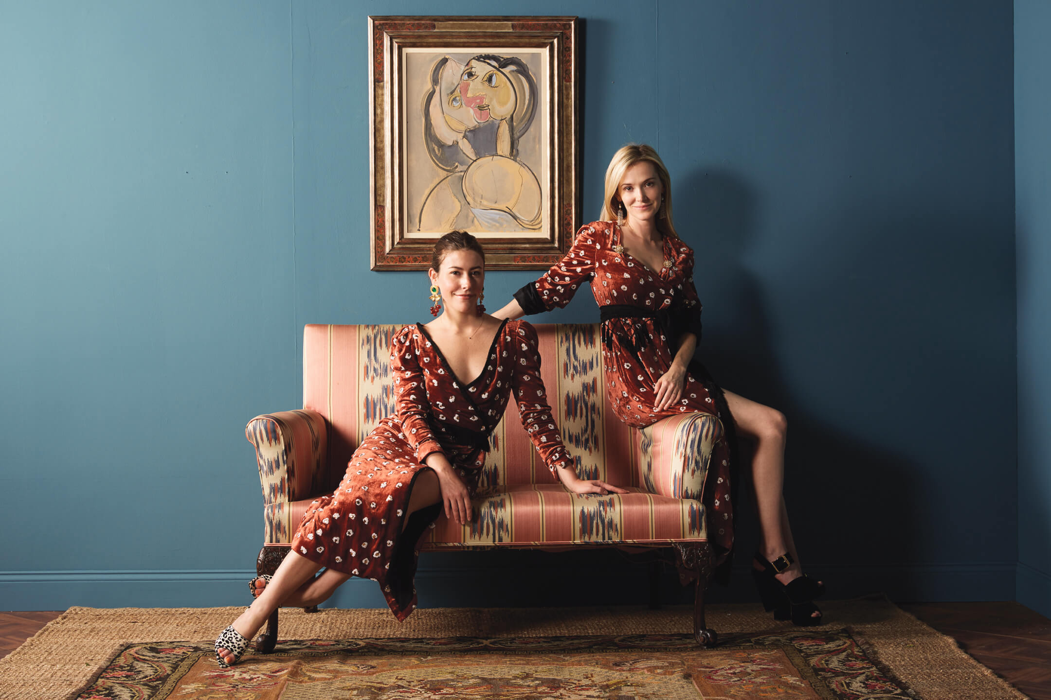 Two women in patterned dresses sitting on a striped sofa, with a framed painting on a blue wall background.