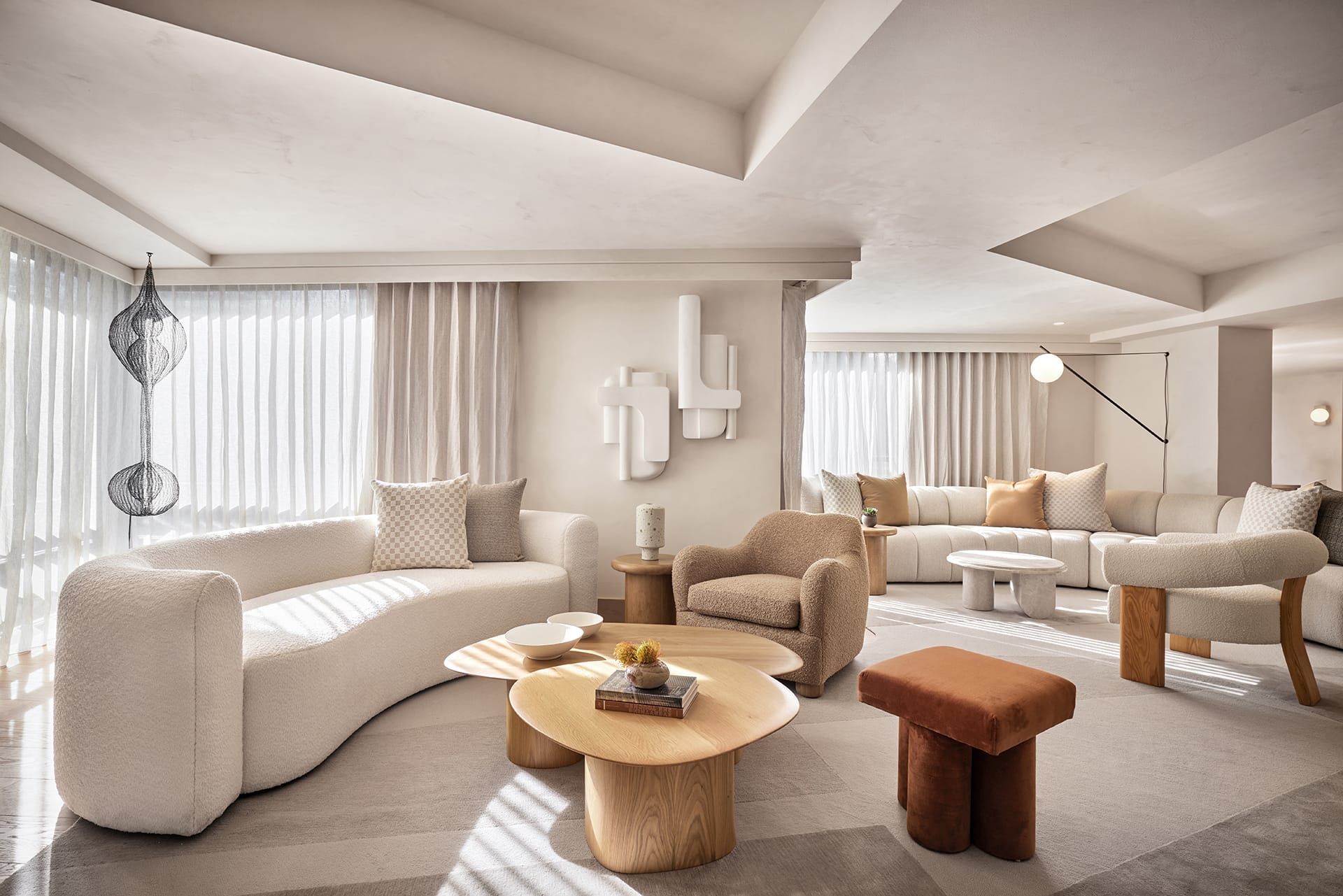 Modern living room with beige curved sofas, round wooden coffee tables, and natural light streaming through sheer curtains.