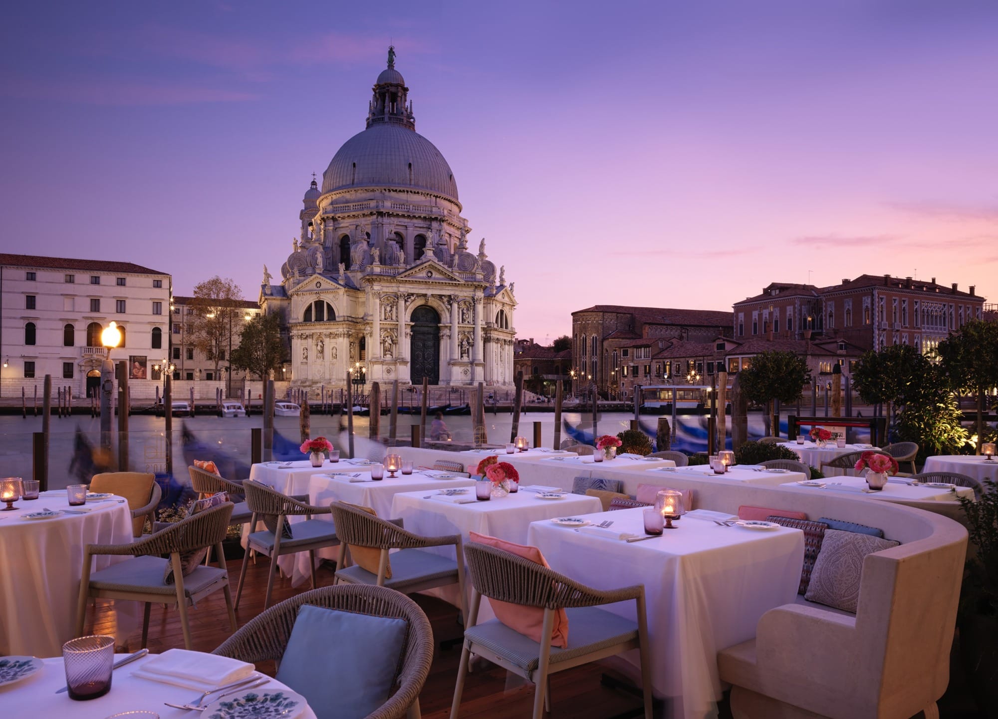 Rooftop restaurant at sunset overlooking a canal and historic domed building in Venice, Italy, with elegantly set tables.