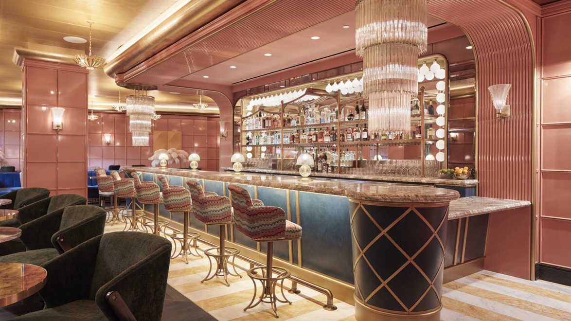 Elegant bar interior with upholstered stools, art deco lighting, and stocked shelves against pink and blue decor.