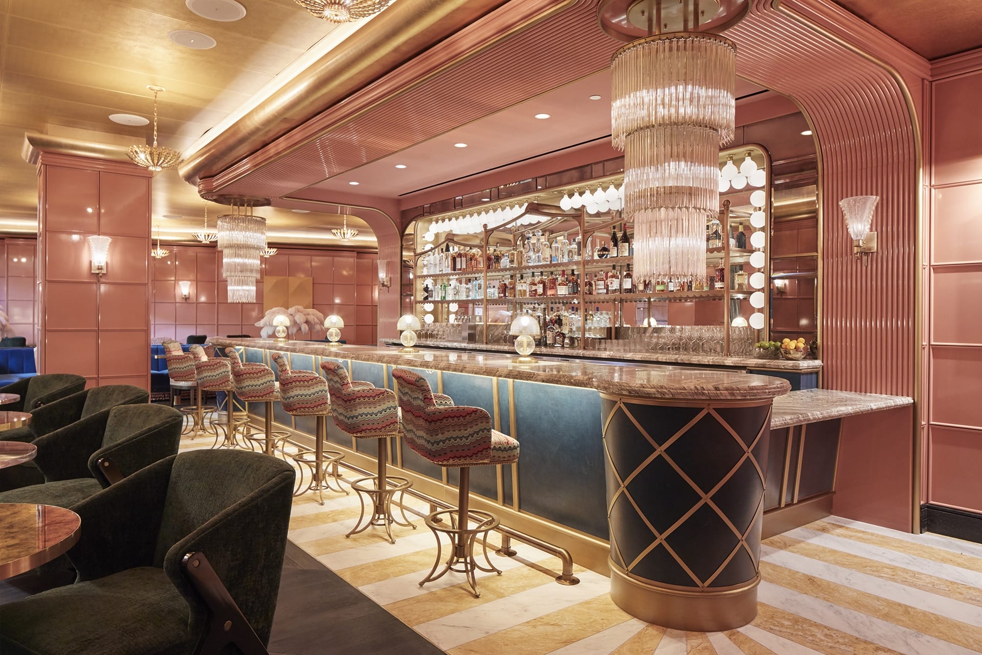 Elegant bar interior with upholstered stools, art deco lighting, and stocked shelves against pink and blue decor.