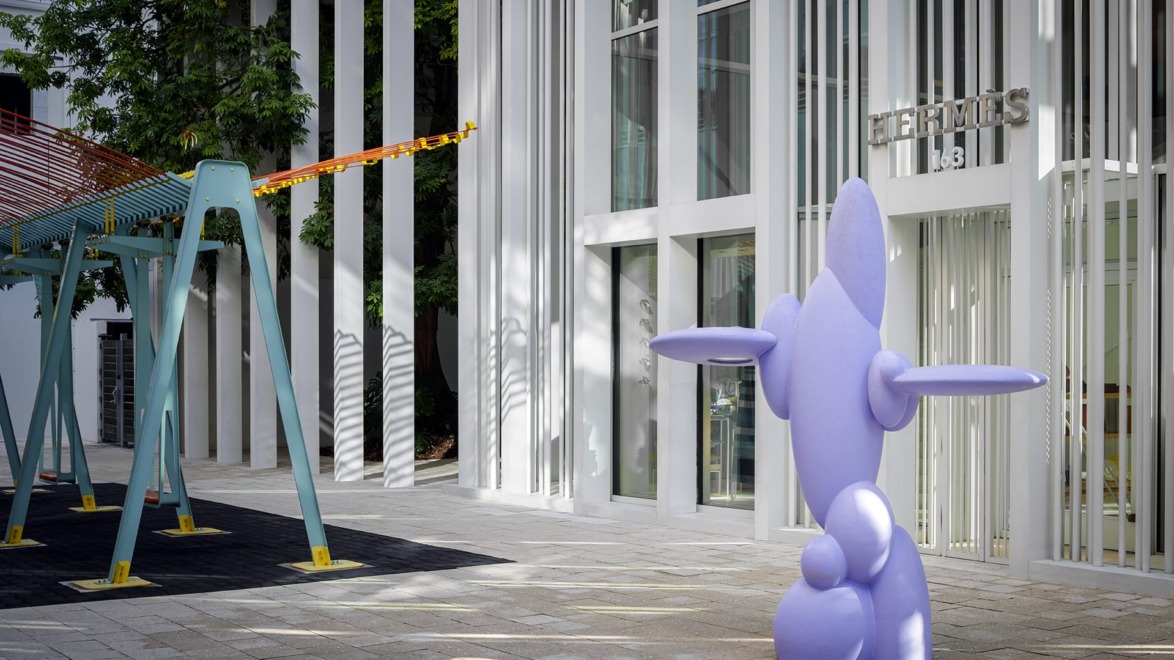 Purple abstract sculpture in front of a modern building with vertical lines and a swing set nearby.