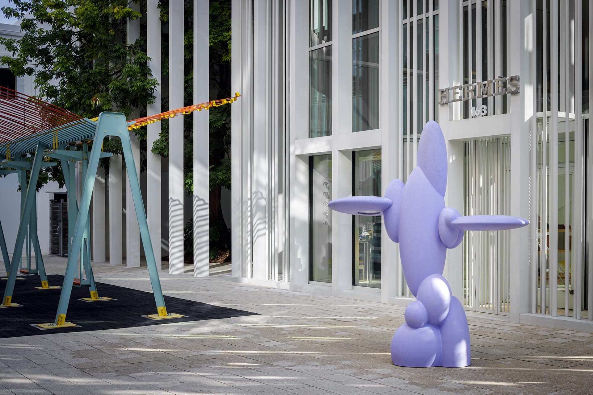 Purple abstract sculpture in front of a modern building with vertical lines and a swing set nearby.