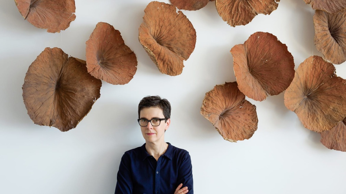 Person standing against a wall with brown, leaf-like sculptures, arms crossed, wearing glasses and a dark shirt.
