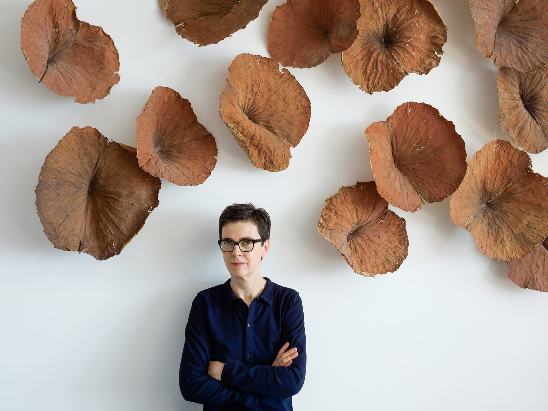 Person standing against a wall with brown, leaf-like sculptures, arms crossed, wearing glasses and a dark shirt.
