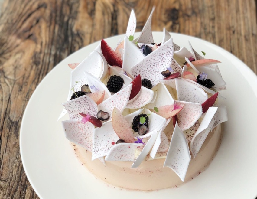Elegant cake with white and pink triangular decorations, berries, and edible flowers on a white plate, wooden table background.