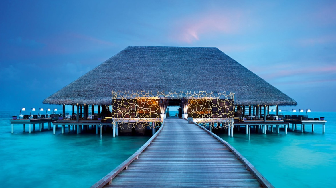 Wooden walkway leading to an overwater bungalow at sunset with a thatched roof and glowing lights, surrounded by calm blue water.