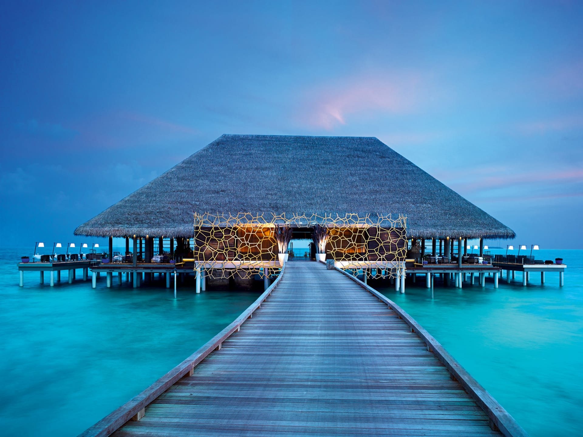 Wooden walkway leading to an overwater bungalow at sunset with a thatched roof and glowing lights, surrounded by calm blue water.